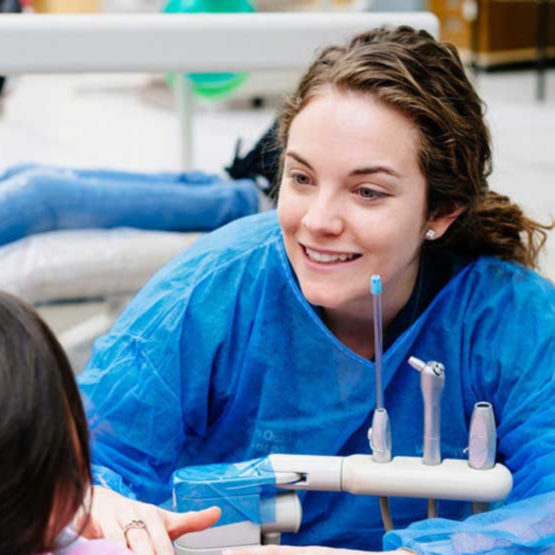 A women with brown hair wearing a blue medical gown smiling at child in dental chair